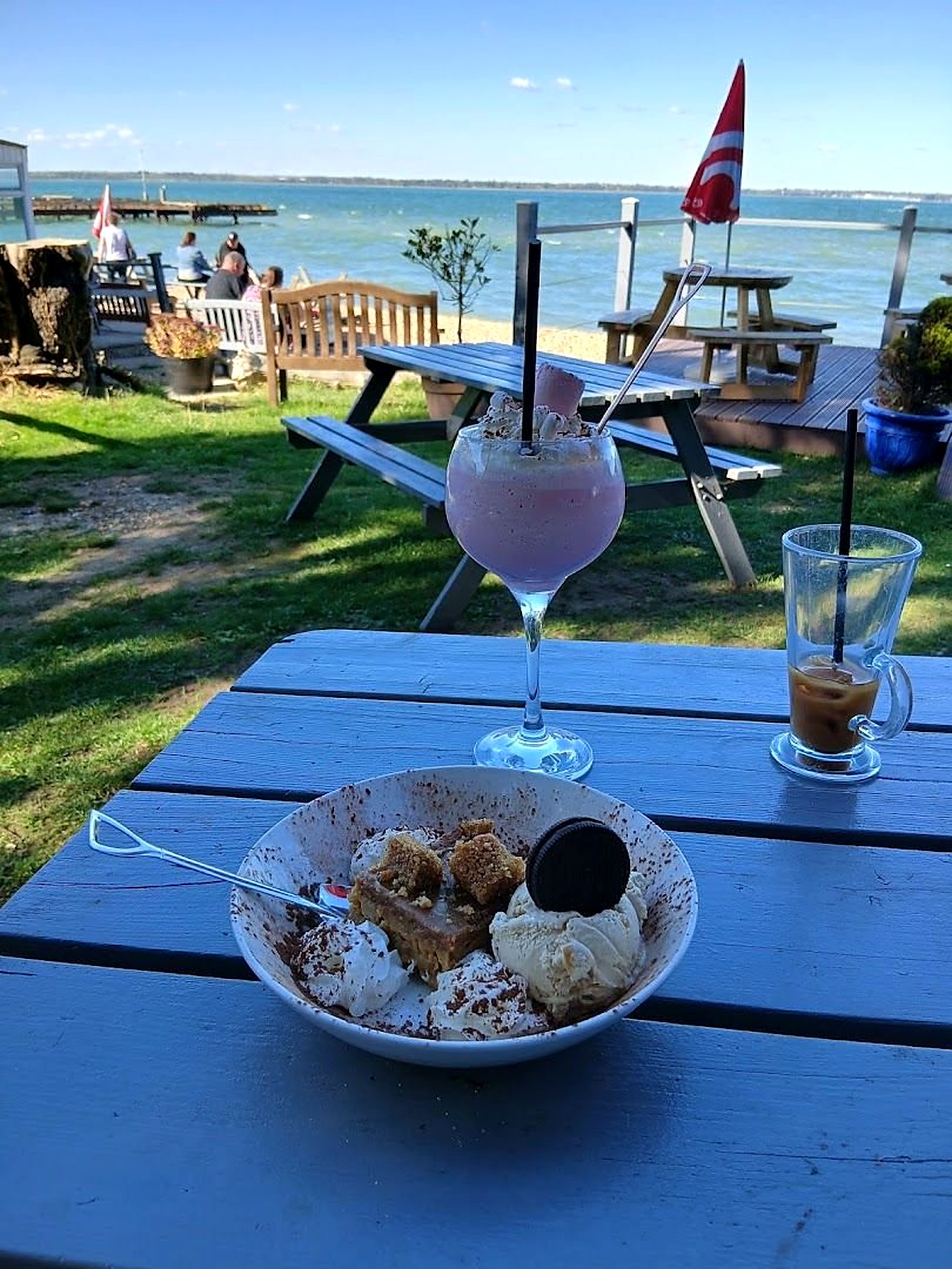A blue picnic table in the Boat House garden with an ice-cream sundae and drinks, the sea and a red flag behind